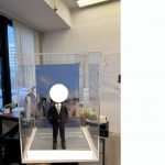 透明展示盒內的西裝主題公仔：身穿深色西裝與領帶的男子雙手插袋站在灰色地台上，背後是藍天白雲下的城市天際線照片背板，前方有可客製銘牌。