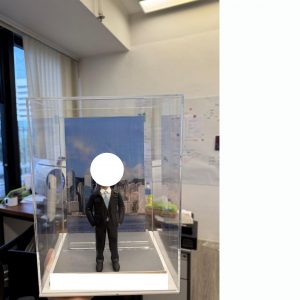透明展示盒內的西裝主題公仔：身穿深色西裝與領帶的男子雙手插袋站在灰色地台上，背後是藍天白雲下的城市天際線照片背板，前方有可客製銘牌。