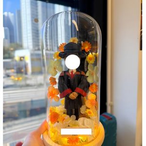 人像畢業公仔穿黑色學袍與方帽,配紅黑披肩,置於木座玻璃罩內,周圍點綴橙黃與白色花材並有暖色燈光,前方附可客製銘牌