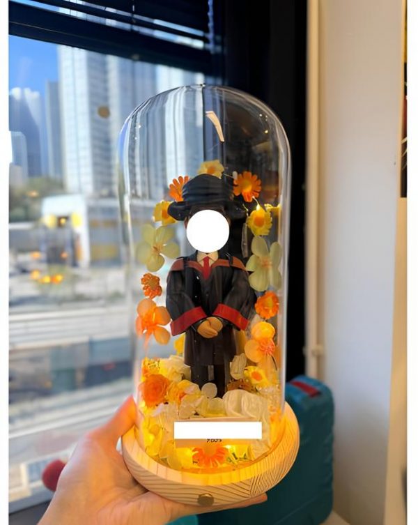 人像畢業公仔穿黑色學袍與方帽，配紅黑披肩，置於木座玻璃罩內，周圍點綴橙黃與白色花材並有暖色燈光，前方附可客製銘牌