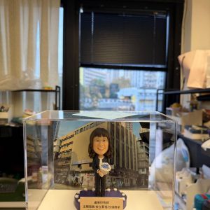 透明展示盒內的客製牧師公仔，女士手捧地球儀站立於紫色底座前，背板為教會建築外觀，前方銘牌刻有祝福與日期