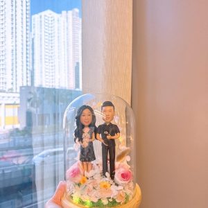玻璃花盅內的一對客製情侶公仔，女方穿深色花裙抱白色小狗，男方穿黑色襯衫抱黑色小狗，周圍以粉玫瑰、白花與暖光燈點綴，木質底座襯草地