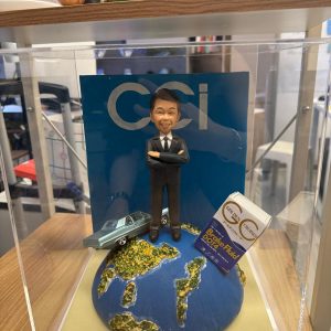 透明展示盒內的客製男士公仔,穿黑色西裝雙手抱胸站在地球造型底座上,旁邊有復古藍綠小汽車與「Brake Fluid DOT4」油品盒,背板印有「CCI」字樣