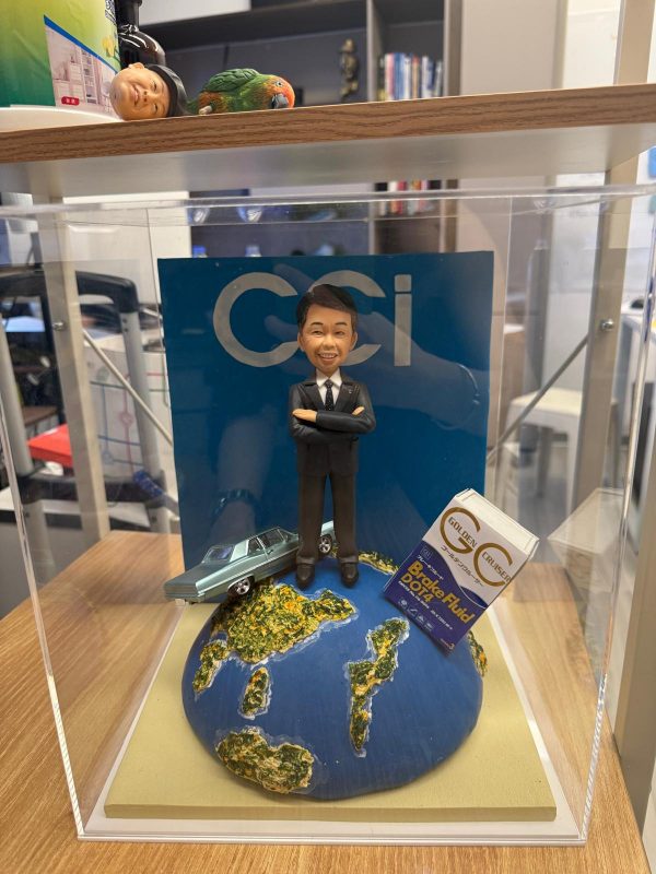 透明展示盒內的客製男士公仔，穿黑色西裝雙手抱胸站在地球造型底座上，旁邊有復古藍綠小汽車與「Brake Fluid DOT4」油品盒，背板印有「CCI」字樣