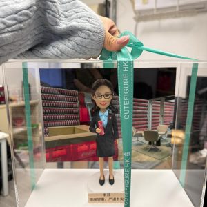 手提綠色緞帶的透明展示盒,內有客製女主管公仔,穿深色套裝配藍色襯衫與領巾,手持可口可樂易拉罐,背板為堆滿可樂的貨架與辦公休息區