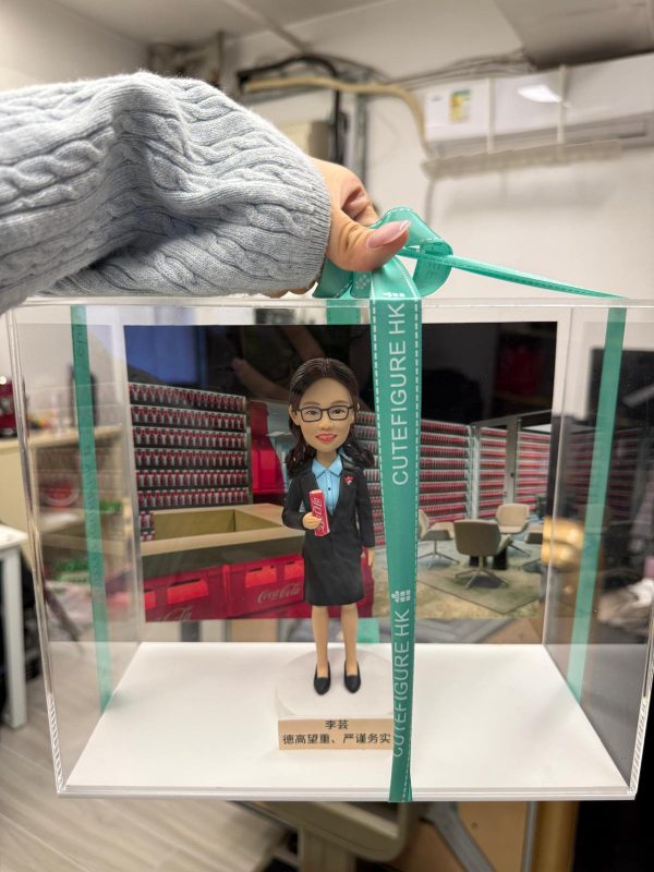 手提綠色緞帶的透明展示盒，內有客製女主管公仔，穿深色套裝配藍色襯衫與領巾，手持可口可樂易拉罐，背板為堆滿可樂的貨架與辦公休息區