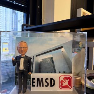 透明展示盒內的客製退休紀念公仔，男士穿黑色西裝打領結，比手勢站在灰色台座上；前方有「EMSD」標誌牌，背板為大樓外觀，銘牌寫著「Yorkie Sir Happy Retirement 2026」