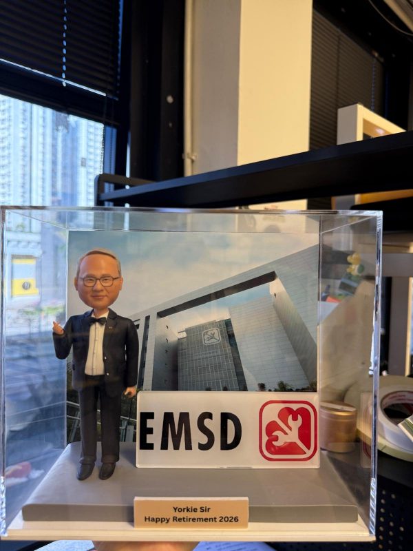 透明展示盒內的客製退休紀念公仔，男士穿黑色西裝打領結，比手勢站在灰色台座上；前方有「EMSD」標誌牌，背板為大樓外觀，銘牌寫著「Yorkie Sir Happy Retirement 2026」