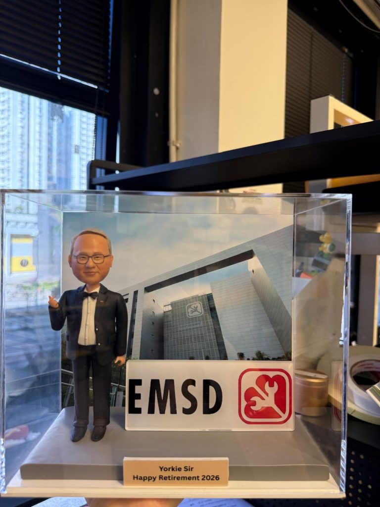 透明展示盒內的客製退休紀念公仔，男士穿黑色西裝打領結，比手勢站在灰色台座上；前方有「EMSD」標誌牌，背板為大樓外觀，銘牌寫著「Yorkie Sir Happy Retirement 2026」