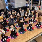 辦公室枱面排放多個客製人像公仔，立於彩色圓形底座並刻上姓名；角色造型多樣，包含黑白格外套、抱公仔、拿籃球與咖啡等個人元素，整體氣氛熱鬧有趣