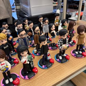 辦公室枱面排放多個客製人像公仔，立於彩色圓形底座並刻上姓名；角色造型多樣，包含黑白格外套、抱公仔、拿籃球與咖啡等個人元素，整體氣氛熱鬧有趣