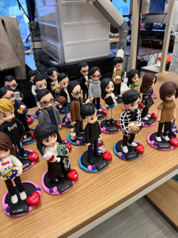 辦公室枱面排放多個客製人像公仔，立於彩色圓形底座並刻上姓名；角色造型多樣，包含黑白格外套、抱公仔、拿籃球與咖啡等個人元素，整體氣氛熱鬧有趣