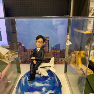 透明展示盒內的客製男士公仔,穿黑色西裝坐在白色小飛機上,飛機停靠於藍白雲紋的地球造型底座前,背板為城市高樓夜景