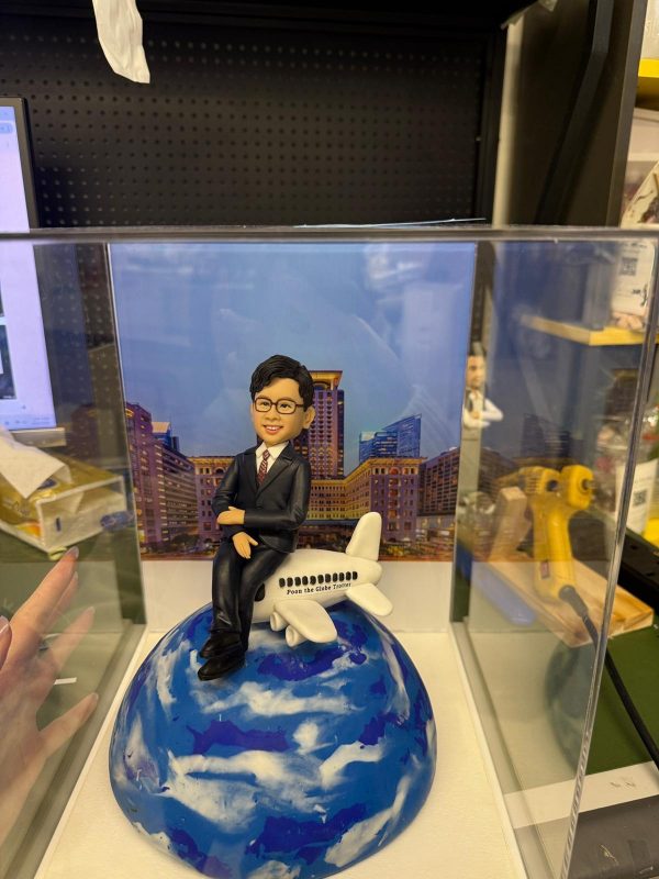 透明展示盒內的客製男士公仔，穿黑色西裝坐在白色小飛機上，飛機停靠於藍白雲紋的地球造型底座前，背板為城市高樓夜景
