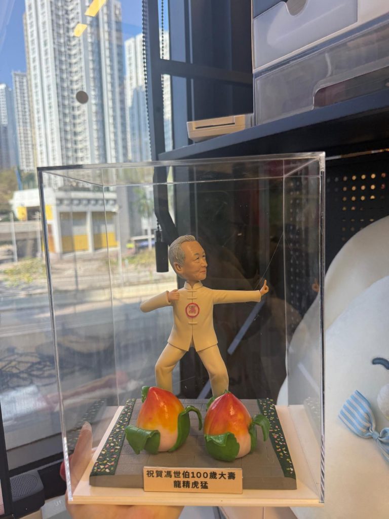 透明展示盒內的客製百歲壽辰公仔，長者穿白色太極服持劍擺姿，前方擺放兩個壽桃與祝賀銘牌，地台為灰色磚面與花紋邊條