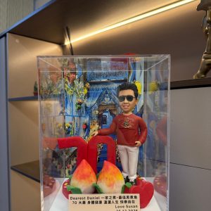 透明展示盒內的客製公仔七十大壽禮物,主角穿紅色上衣與白褲、戴墨鏡,背景為藍白小鎮拱門場景,前方有紅色「70」與兩個壽桃及心形裝飾,附祝福銘牌