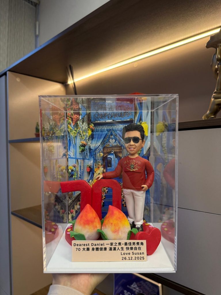 透明展示盒內的客製公仔七十大壽禮物，主角穿紅色上衣與白褲、戴墨鏡，背景為藍白小鎮拱門場景，前方有紅色「70」與兩個壽桃及心形裝飾，附祝福銘牌