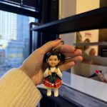 手掌捏著客製娃娃吊飾，女孩綁雙馬尾，穿白色上衣搭綠紅相間的聖誕風吊帶裙與毛球裝飾，表情可愛俏皮