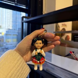 手掌捏著客製娃娃吊飾,女孩綁雙馬尾,穿白色上衣搭綠紅相間的聖誕風吊帶裙與毛球裝飾,表情可愛俏皮