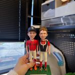 手掌托著雙人少棒公仔，兩位男孩穿紅黑配色球衣與白色球褲並肩站立，左側戴深藍帽、右側穿護胸背心，綠色草地底座上有名牌「Brady & Ryan」