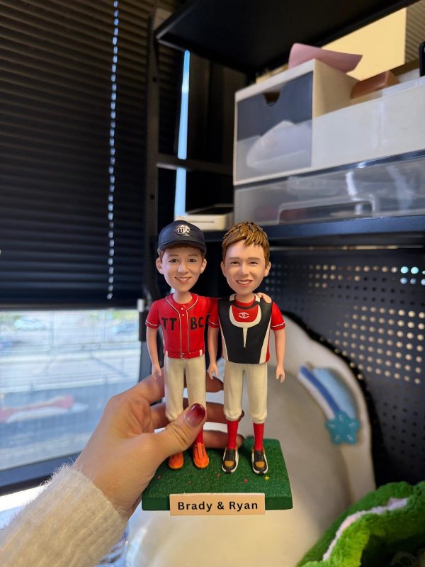 手掌托著雙人少棒公仔，兩位男孩穿紅黑配色球衣與白色球褲並肩站立，左側戴深藍帽、右側穿護胸背心，綠色草地底座上有名牌「Brady & Ryan」