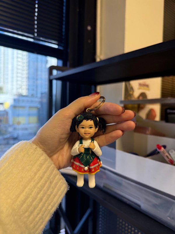 手掌捏著客製娃娃吊飾，女孩綁雙馬尾，穿白色上衣搭綠紅相間的聖誕風吊帶裙與毛球裝飾，表情可愛俏皮