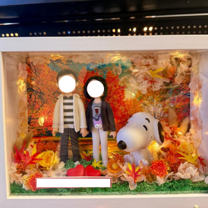 白色相框燈箱內的客製情侶公仔場景,背景為秋日楓葉林,周圍以乾燥花與暖黃小燈點綴,前方擺放兩顆紅心與可愛史努比模型