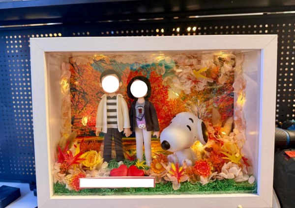 白色相框燈箱內的客製情侶公仔場景，背景為秋日楓葉林，周圍以乾燥花與暖黃小燈點綴，前方擺放兩顆紅心與可愛史努比模型
