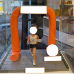 透明展示盒內的客製跑者公仔，穿背心短褲與號碼布，衝過橙色拱門終點線，地面為灰色磚紋跑道並設有銘牌位置