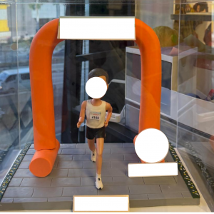 透明展示盒內的客製跑者公仔,穿背心短褲與號碼布,衝過橙色拱門終點線,地面為灰色磚紋跑道並設有銘牌位置