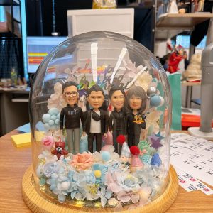 玻璃穹頂裡的四人客製公仔生日花盅，人物穿休閒與黑色外套並肩站立，周圍點綴藍粉永生花、派對帽與生日小物，木質底座溫暖質感