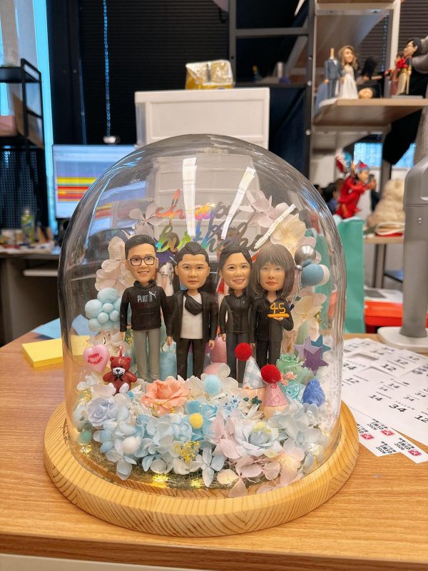 玻璃穹頂裡的四人客製公仔生日花盅，人物穿休閒與黑色外套並肩站立，周圍點綴藍粉永生花、派對帽與生日小物，木質底座溫暖質感