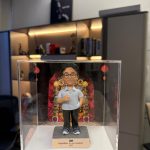 手托透明展示盒，內有客製男士公仔穿淺藍短袖襯衫與黑褲、比讚站姿，背板為喜慶花牌裝飾，底座銘牌寫著「Congratulations on your promotion! 2025」