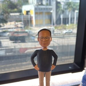 手掌托著客製男士公仔，黑色上衣配卡其長褲與白鞋，雙手插腰站在灰色石紋底座上，作為送給老闆的禮物