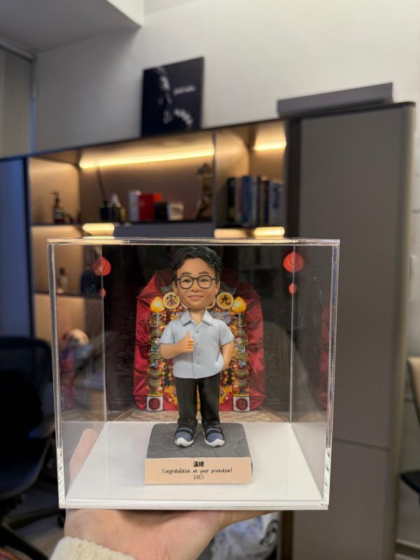 手托透明展示盒，內有客製男士公仔穿淺藍短袖襯衫與黑褲、比讚站姿，背板為喜慶花牌裝飾，底座銘牌寫著「Congratulations on your promotion! 2025」