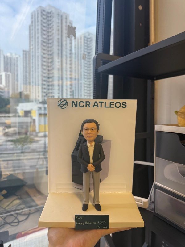 手托透明展示台上的客製退休紀念公仔，男士穿西裝站姿端正，背板印有「NCR ATLEOS」與設備圖，前方銘牌寫著「Keith Happy Retirement 2025 TW Team」