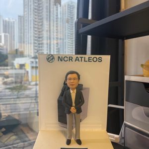 透明展示台上的客製退休公仔，男士穿深色西裝與淺色長褲端正站立，背板印有「NCR ATLEOS」與設備圖，前方綠色銘牌寫「Keith Happy Retirement 2025 TW Team」