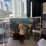 透明展示盒內的客製退休公仔，男士穿西裝微笑持證書，身後有兩隻海豚與圓環裝飾，背板為海岸與校園俯瞰圖，前方銘牌寫「Raymond sir Happy Retirement 2026」