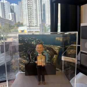 透明展示盒內的客製退休公仔，男士穿西裝微笑持證書，身後有兩隻海豚與圓環裝飾，背板為海岸與校園俯瞰圖，前方銘牌寫「Raymond sir Happy Retirement 2026」