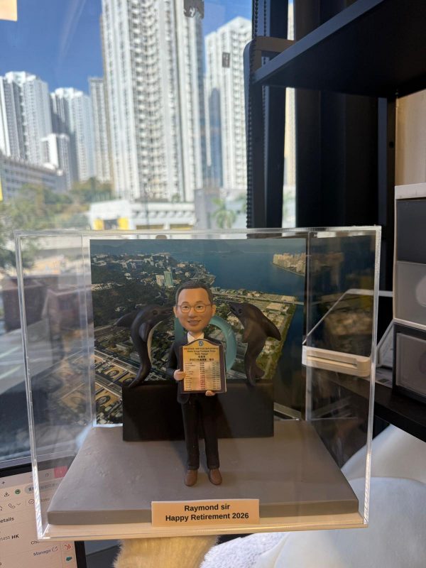 透明展示盒內的客製退休公仔，男士穿西裝微笑持證書，身後有兩隻海豚與圓環裝飾，背板為海岸與校園俯瞰圖，前方銘牌寫「Raymond sir Happy Retirement 2026」