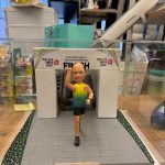 透明展示盒中的客製退休公仔，男士穿漸層跑步背心與短褲、衝線舉手慶祝，身後是印有「EMSD 2026」與 FINISH 的終點拱門，前方銘牌寫著「Happy Retirement to PC Sir」