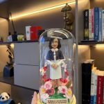 玻璃花盅內的客製女護士公仔，穿白色制服披深藍披肩，周圍以粉紅玫瑰與小花環繞並有暖黃色小燈，木質底座附祝福銘牌