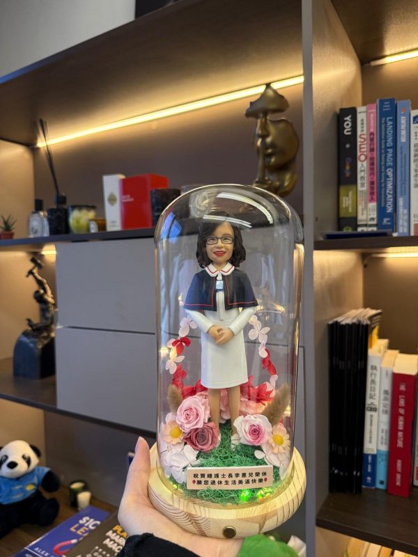 玻璃花盅內的客製女護士公仔，穿白色制服披深藍披肩，周圍以粉紅玫瑰與小花環繞並有暖黃色小燈，木質底座附祝福銘牌