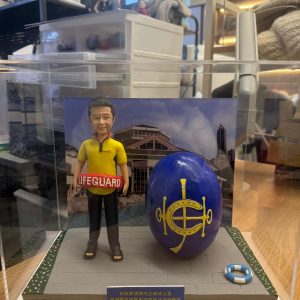 透明展示盒中的救生員主題公仔,穿黃黑配色上衣與長褲、腳穿涼鞋,腰間掛著紅色「LIFEGUARD」浮條;旁邊有一顆大型藍色橢圓物,上有金色標誌;腳下為灰色地磚地台,邊緣綠地點綴小花,右前方放著迷你救生圈;背板是泳池與建築的照片;前方藍底金字名牌寫有祝賀與日期。