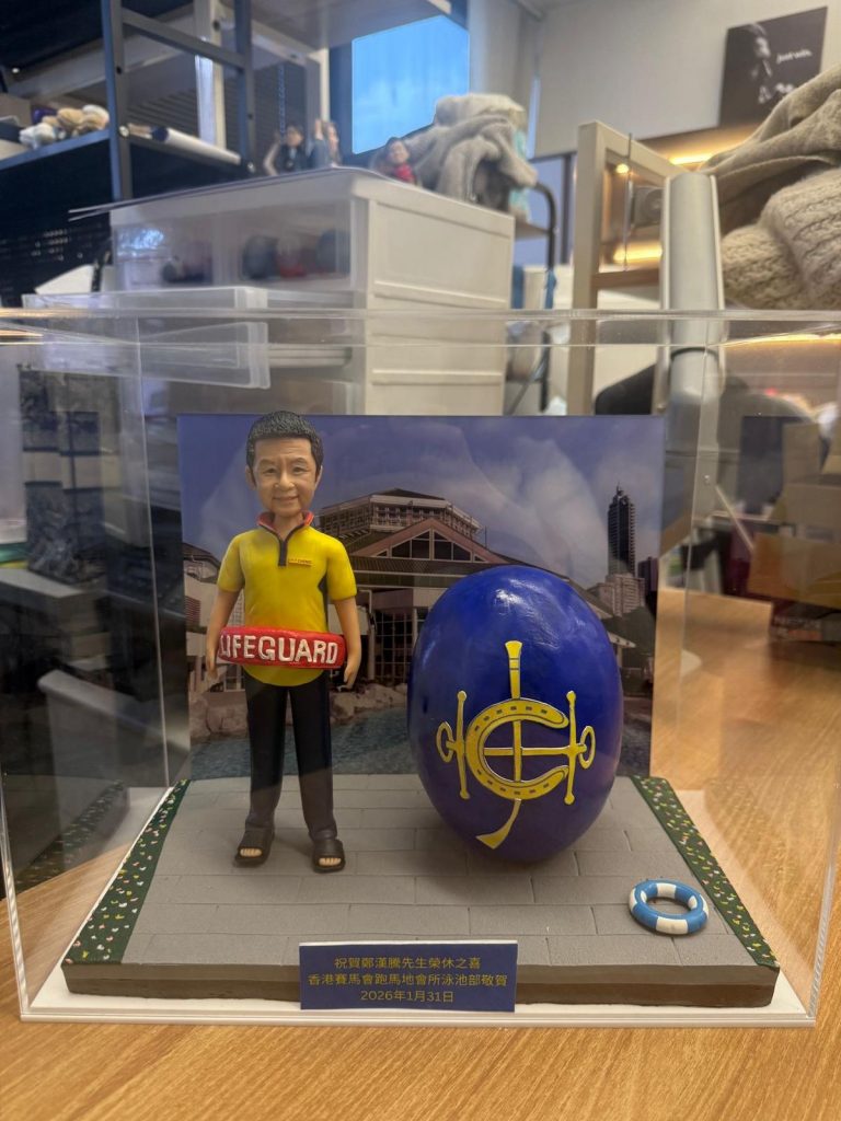 透明展示盒中的救生員主題公仔，穿黃黑配色上衣與長褲、腳穿涼鞋，腰間掛著紅色「LIFEGUARD」浮條；旁邊有一顆大型藍色橢圓物，上有金色標誌；腳下為灰色地磚地台，邊緣綠地點綴小花，右前方放著迷你救生圈；背板是泳池與建築的照片；前方藍底金字名牌寫有祝賀與日期。