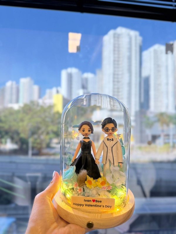 手持玻璃罩小夜燈式雙人公仔：木質圓形底座上放置透明玻璃罩，內有穿黑色禮服與白色西裝的公仔牽手站立，周圍點綴藍色與白色乾燥花、黃色小花與暖黃燈串；前方小名牌寫著「Ivan ♥ Bee Happy Valentine’s Day」。背景為窗外藍天與高樓城市景。