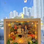 手持白色立體相框式展示盒，內有一家三口公仔：大人兩位與小孩一位，穿著黑白條紋與淺色服裝，前方鋪綠草與淺色花材；四周與背板佈滿多個可愛卡通角色與小擺件，盒內繞有暖黃燈串；前方小牌寫著「Happy Valentine’s Day」。背景是窗外藍天與高樓。