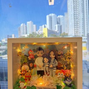 手持白色立體相框式展示盒，內有一家三口公仔：大人兩位與小孩一位，穿著黑白條紋與淺色服裝，前方鋪綠草與淺色花材；四周與背板佈滿多個可愛卡通角色與小擺件，盒內繞有暖黃燈串；前方小牌寫著「Happy Valentine’s Day」。背景是窗外藍天與高樓。