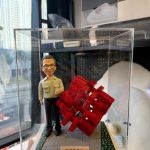 透明展示盒中的客製公仔站在灰色地台上，身穿淺色襯衫與黑色長褲，右手比讚；旁邊有一個紅色編織感的幾何立方體雕塑；地台前方有金色小名牌刻有中文職稱與姓名，四周點綴綠色草地與小花紋路；背景為室內辦公與窗景。