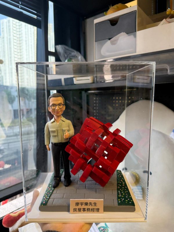 透明展示盒中的客製公仔站在灰色地台上，身穿淺色襯衫與黑色長褲，右手比讚；旁邊有一個紅色編織感的幾何立方體雕塑；地台前方有金色小名牌刻有中文職稱與姓名，四周點綴綠色草地與小花紋路；背景為室內辦公與窗景。
